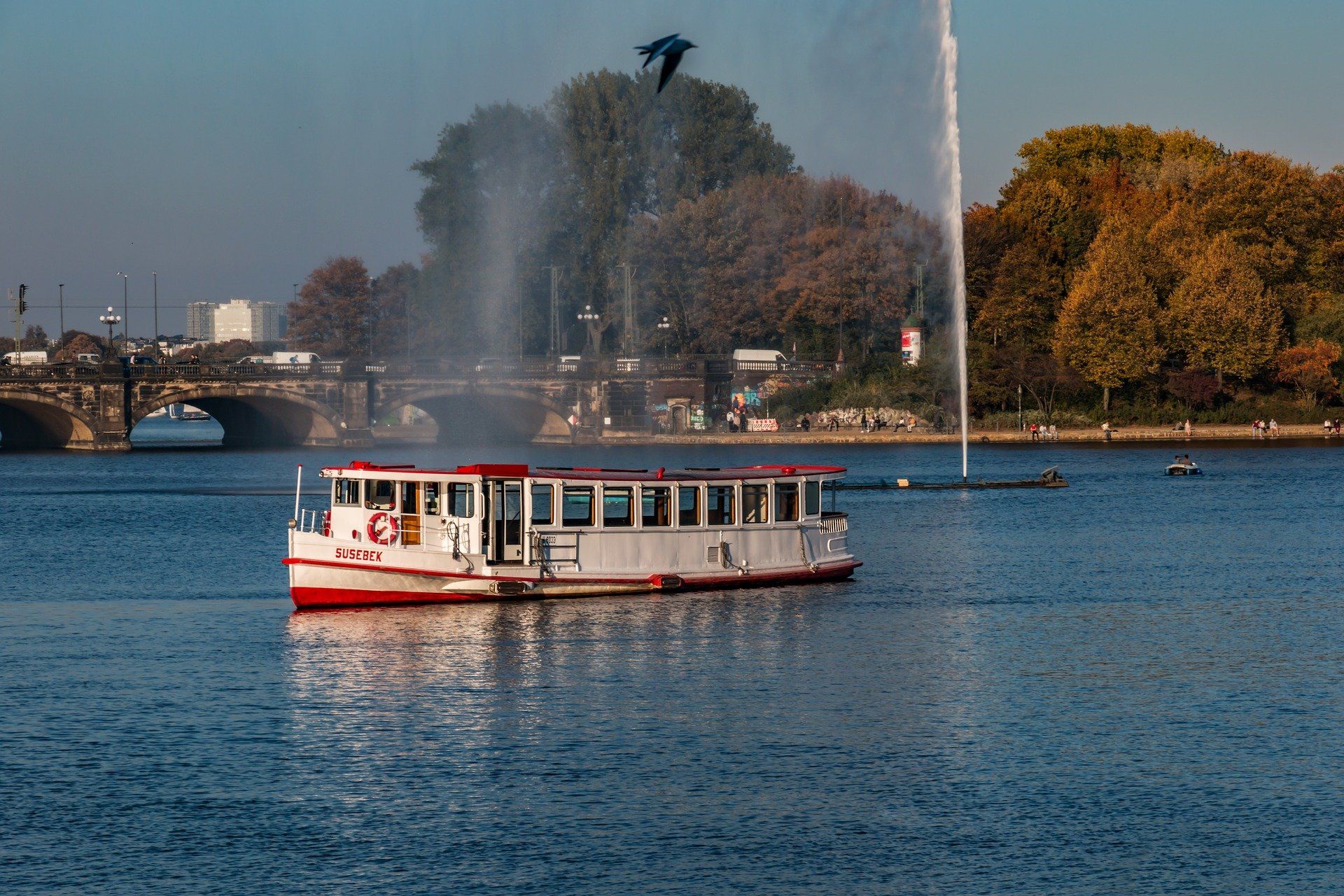 Weißer Alsterdampfer auf blauem Wasser mit Fontänen und Herbstbäumen im Hintergrund