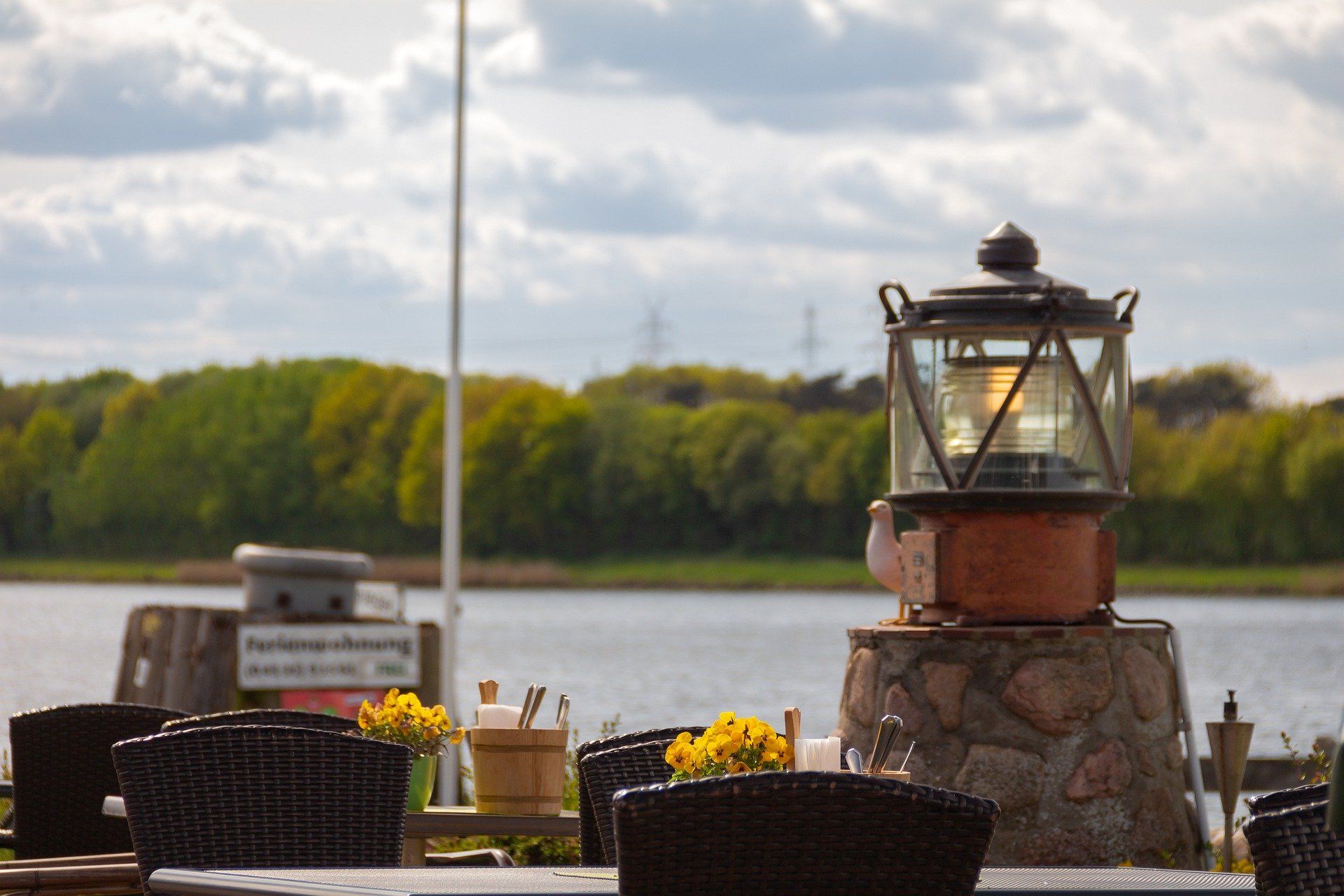 Terrasse am Kanal mit Laterne, Blumen und Korbstühlen mit Blick auf Wasser und Bäume