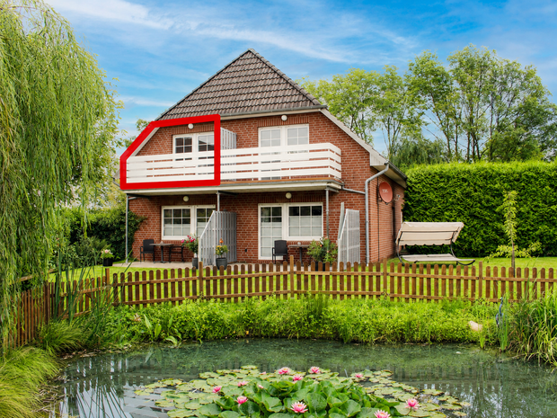 Rotes Backsteinhaus mit weißem Balkon, davor ein Teich und ein Holzzaun in einem grünen Garten