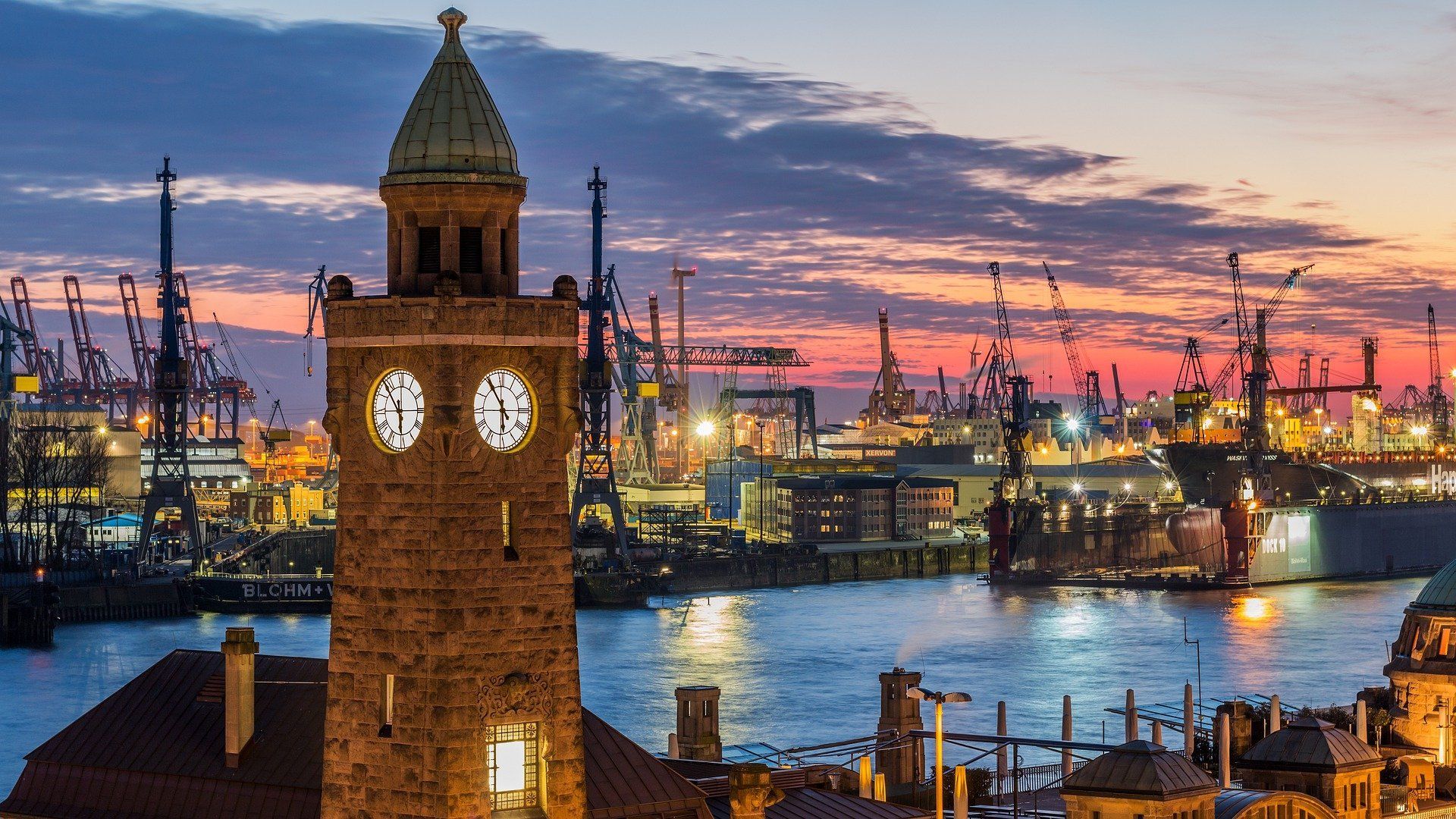 Historischer Glockenturm mit Blick auf einen Hafen bei Sonnenuntergang, im Hintergrund Stadtlichter und Schiffe.