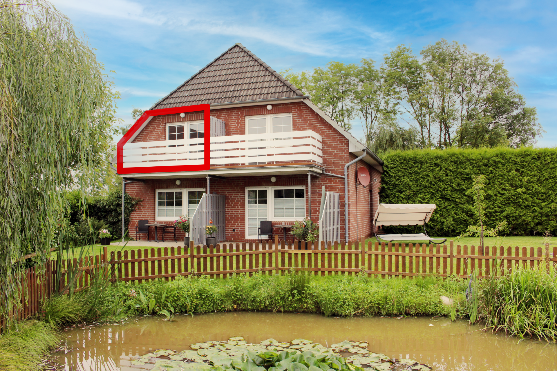 Backsteinhaus mit weißem Balkon mit Blick auf einen Teich und einen von roten Latten umzäunten Garten