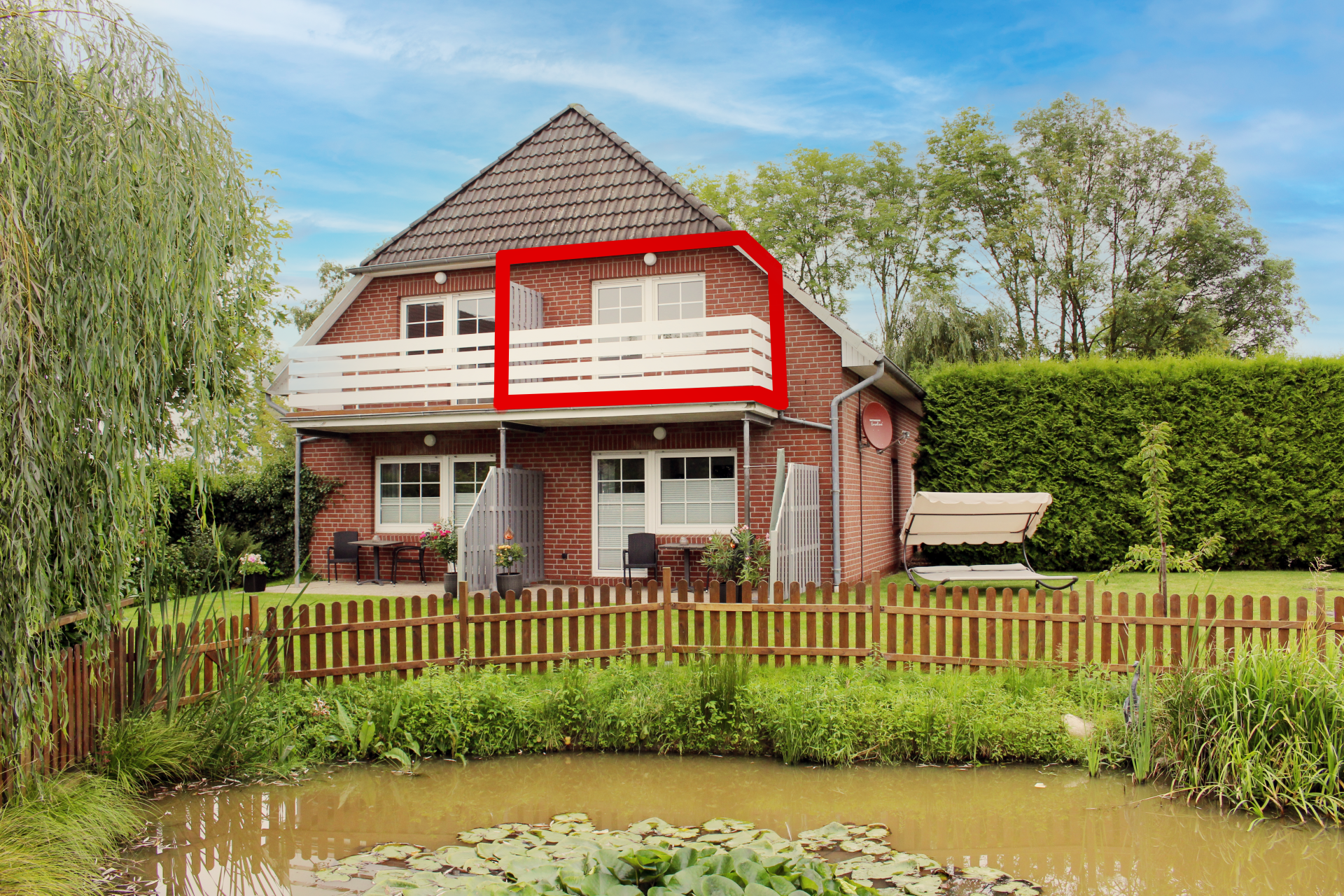 Haus an einem Teich mit einem rot umrandeten Balkon im Obergeschoss, umgeben von Bäumen und einem Holzzaun