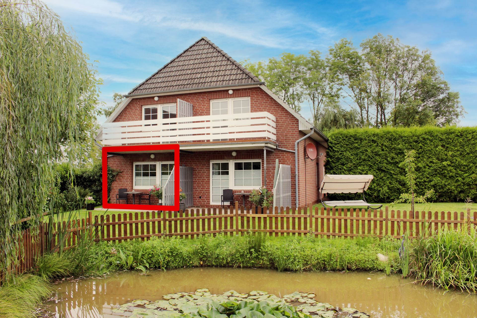 Backsteinhaus mit rot umrandeter Veranda an einem Teich, umgeben von Bäumen und einem Holzzaun