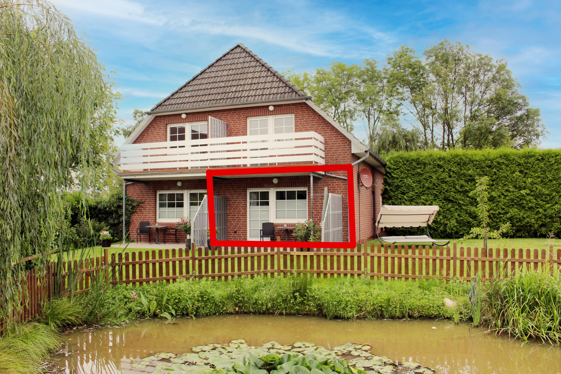 Backsteinhaus mit rotgerahmter Veranda an einem Teich und Holzzaun, umgeben von Bäumen und Grün