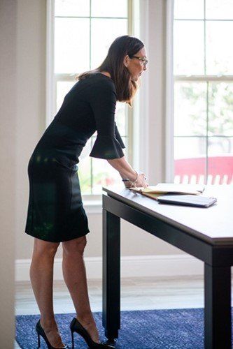 A woman in a black dress is standing next to a desk in front of a window.