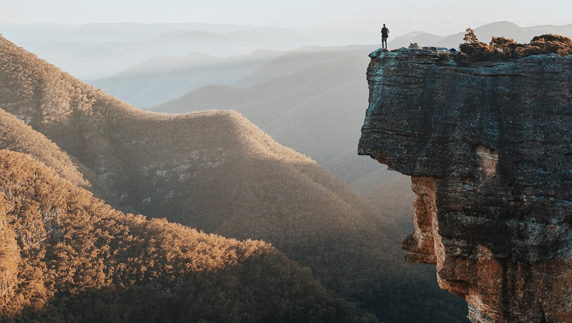 A person is standing on the edge of a cliff overlooking a mountain range.