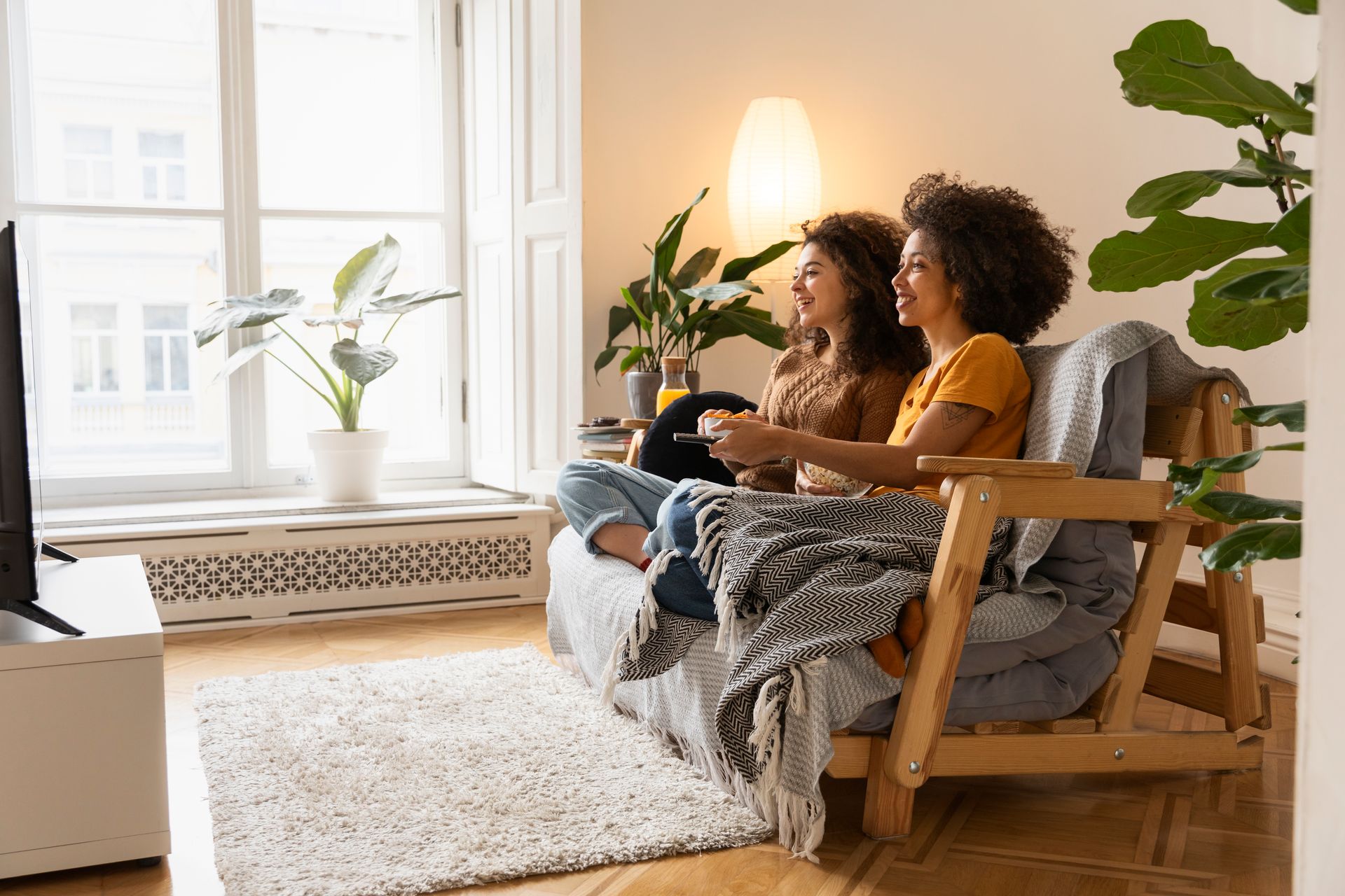 Two people sit on a couch in a bright living room with a plant, lamp, and large window.