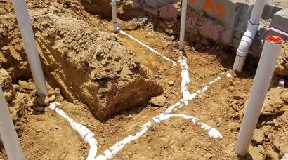 Excavation site with a dirt trench, white PVC pipes, and marked ground lines near a wall
