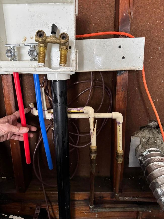 Close-up of exposed plumbing valves and pipes under a sink, with a hand holding a red pipe