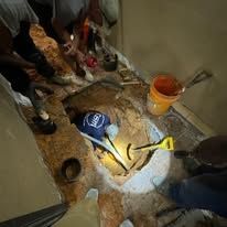 Workers inspecting a damaged floor pipe with flashlight and tools in a dim indoor area