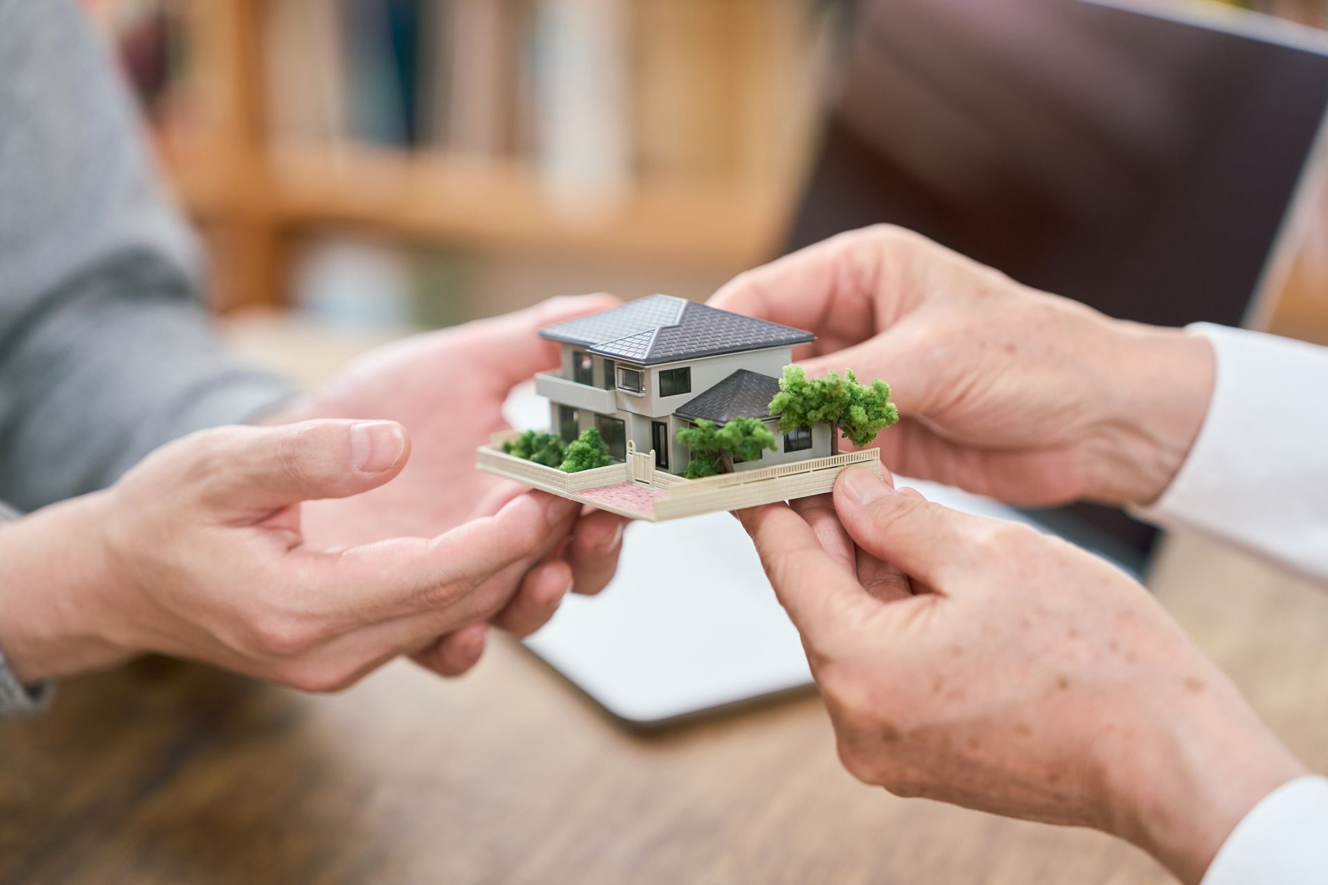 Hands holding a small model house over a wooden table in a bright indoor setting