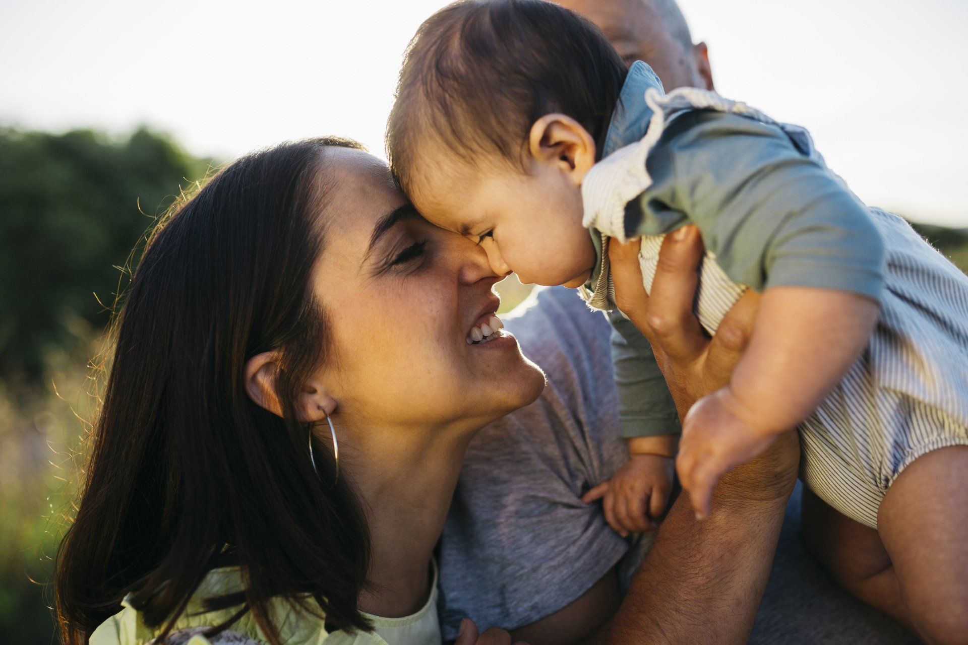 Woman smiling, touching noses with baby held by unseen person outdoors; sunlight.