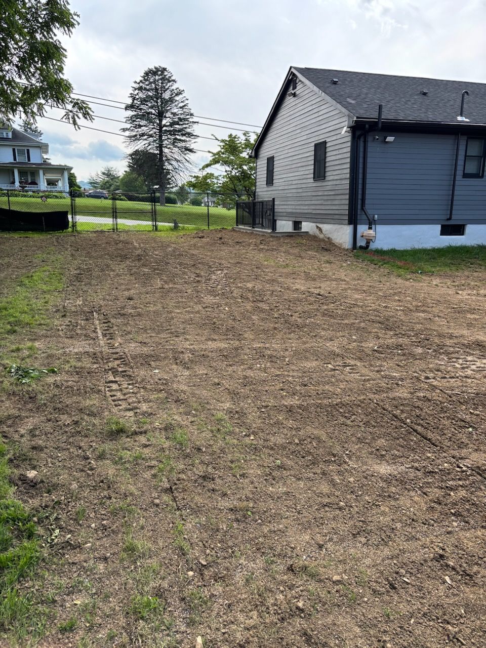 Cleared dirt lot next to a house; grass in foreground, trees and fence in background under a cloudy sky.