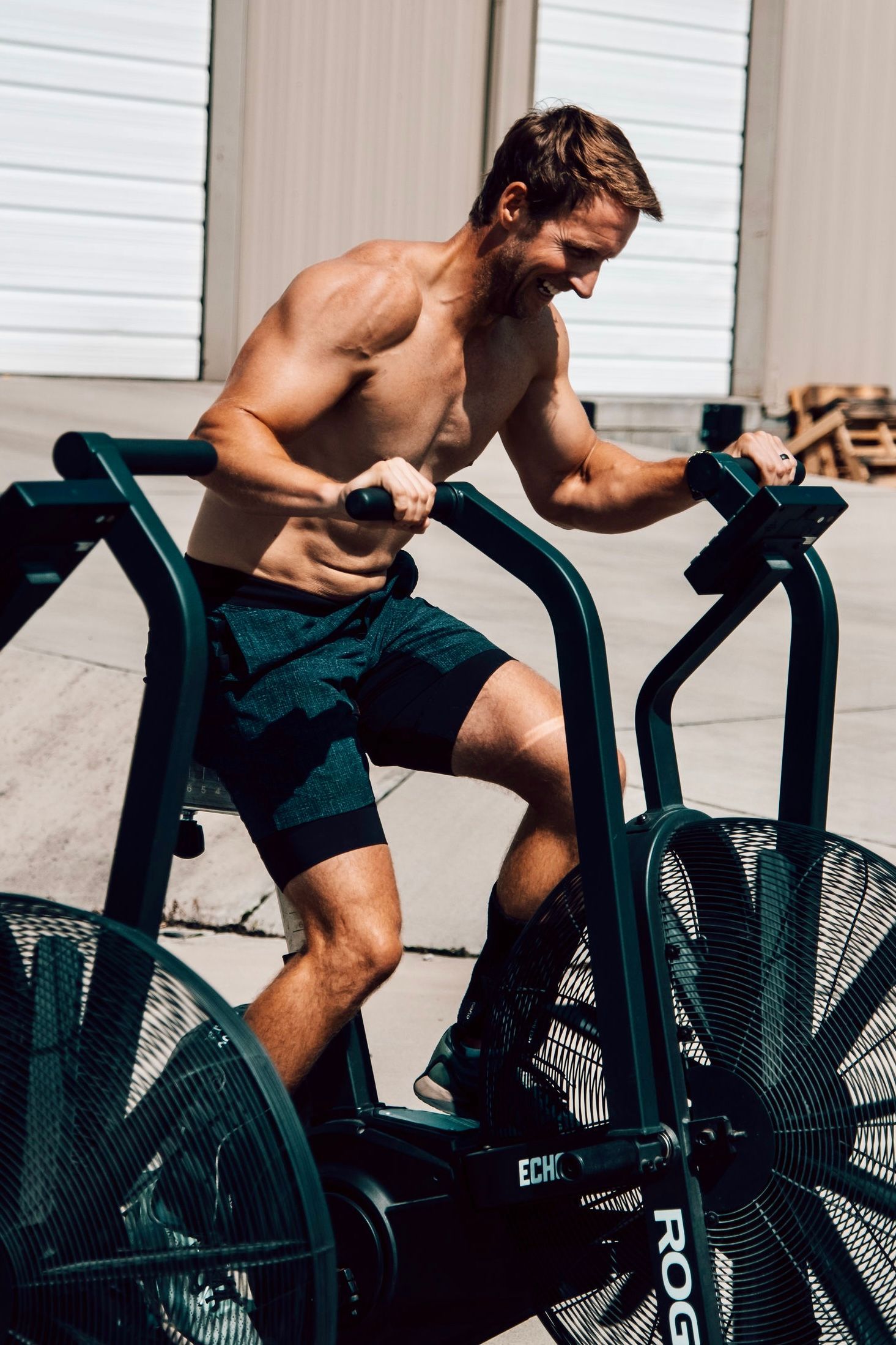 Man exercising on an air bike outdoors in bright sunlight, shirtless and pedaling hard.