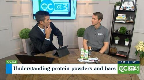 Two men are sitting at a table talking about protein powders and bars.