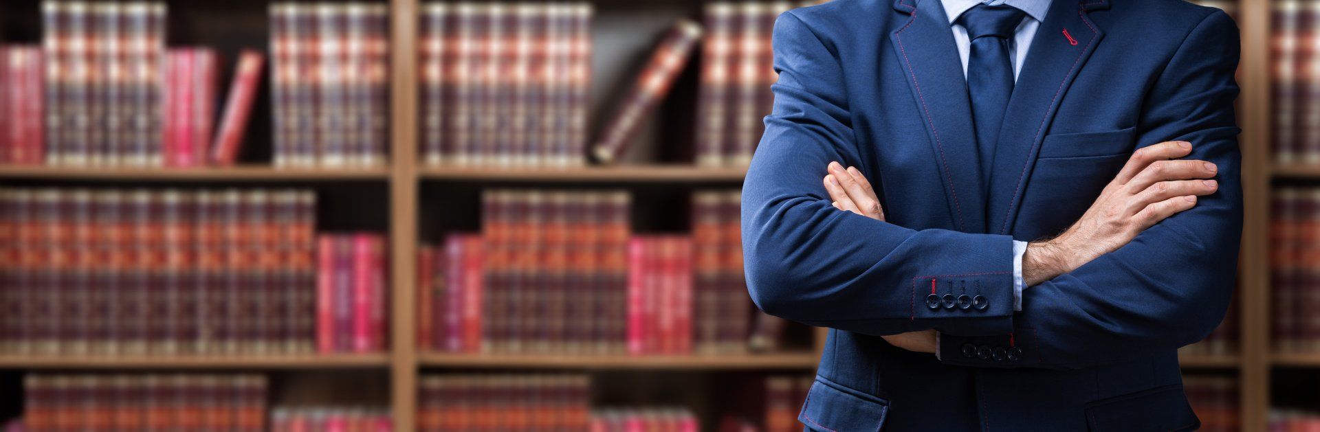 Ein Mann in Anzug und Krawatte steht mit verschränkten Armen vor einem Bücherregal.