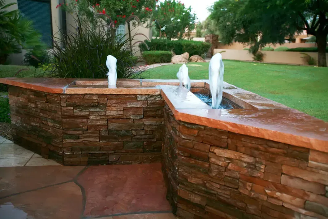 Stone water fountain with three spouts, brown stone wall, green lawn.