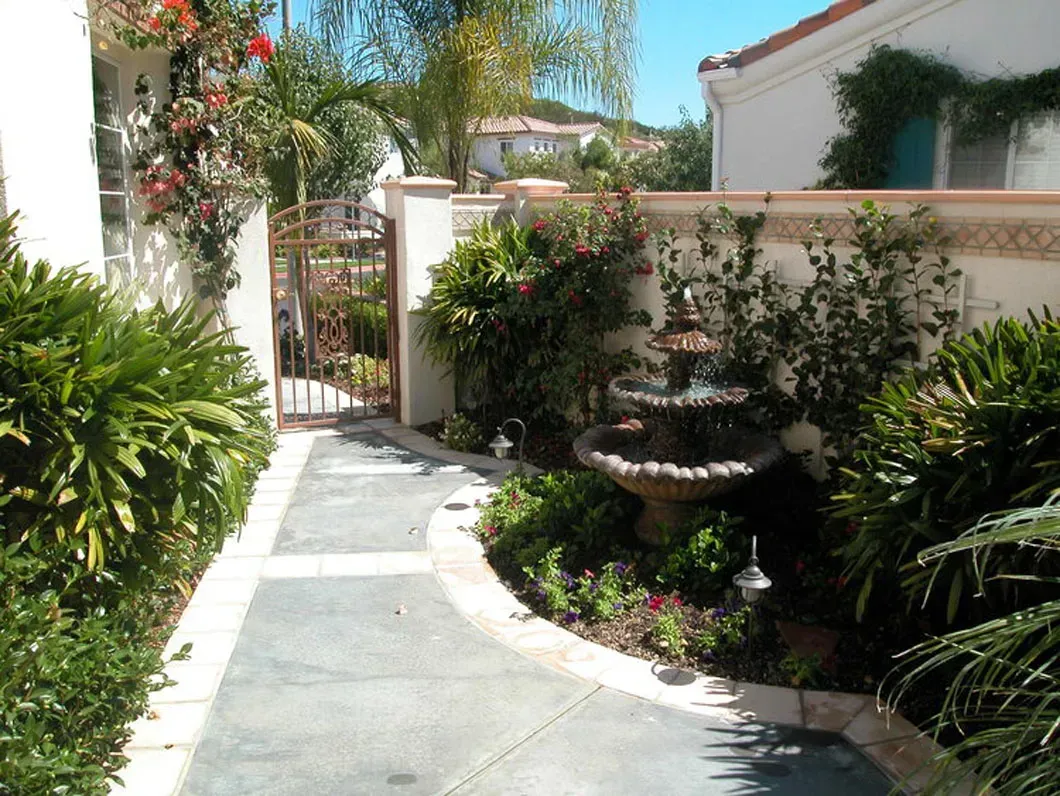 Pathway leading to a wrought iron gate, with a fountain and lush greenery.