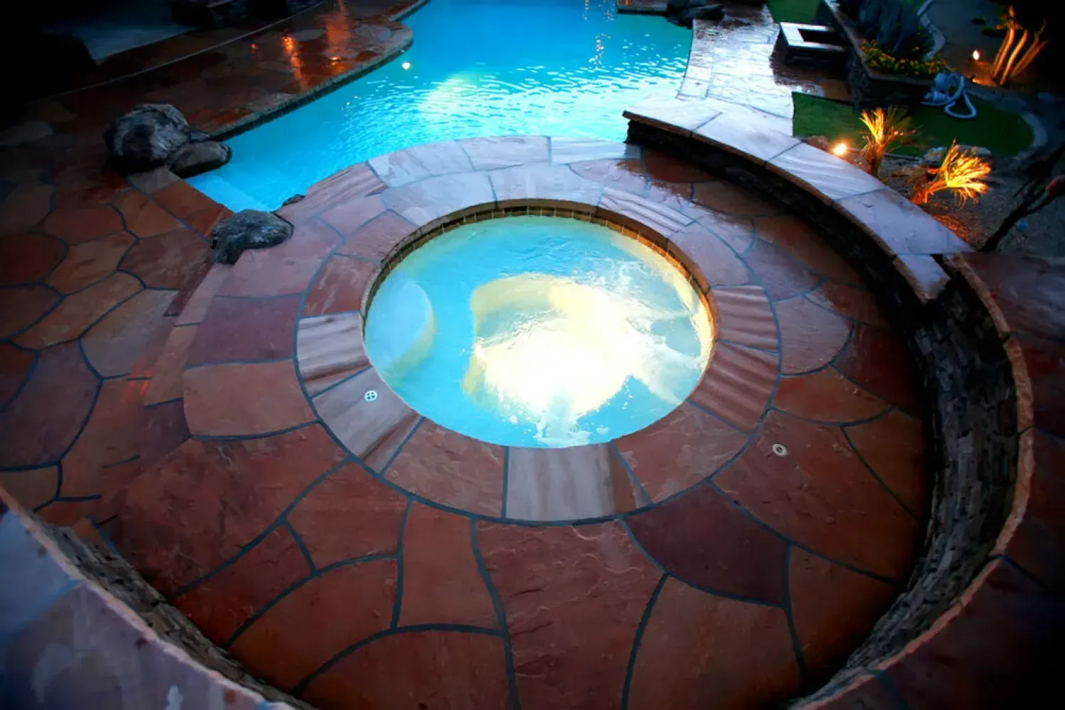 A circular hot tub with water bubbling, surrounded by stone pavers, adjacent to a pool.