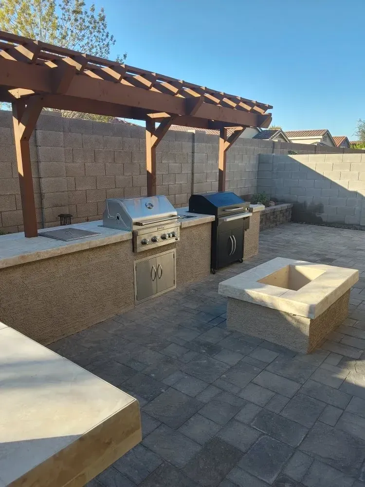 Outdoor kitchen with grill, pergola, and fire pit in a backyard with stone pavers.