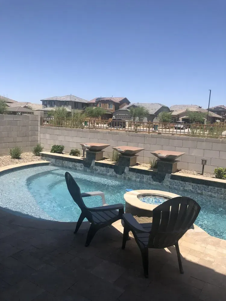 Backyard pool with waterfalls, two chairs, and a fire pit, with houses in the background.