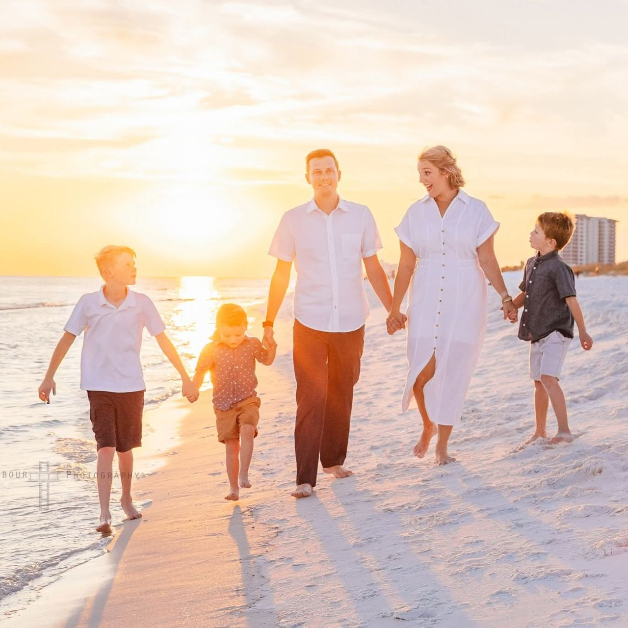 A family walking hand-in-hand along a sandy beach during a bright, golden sunset.