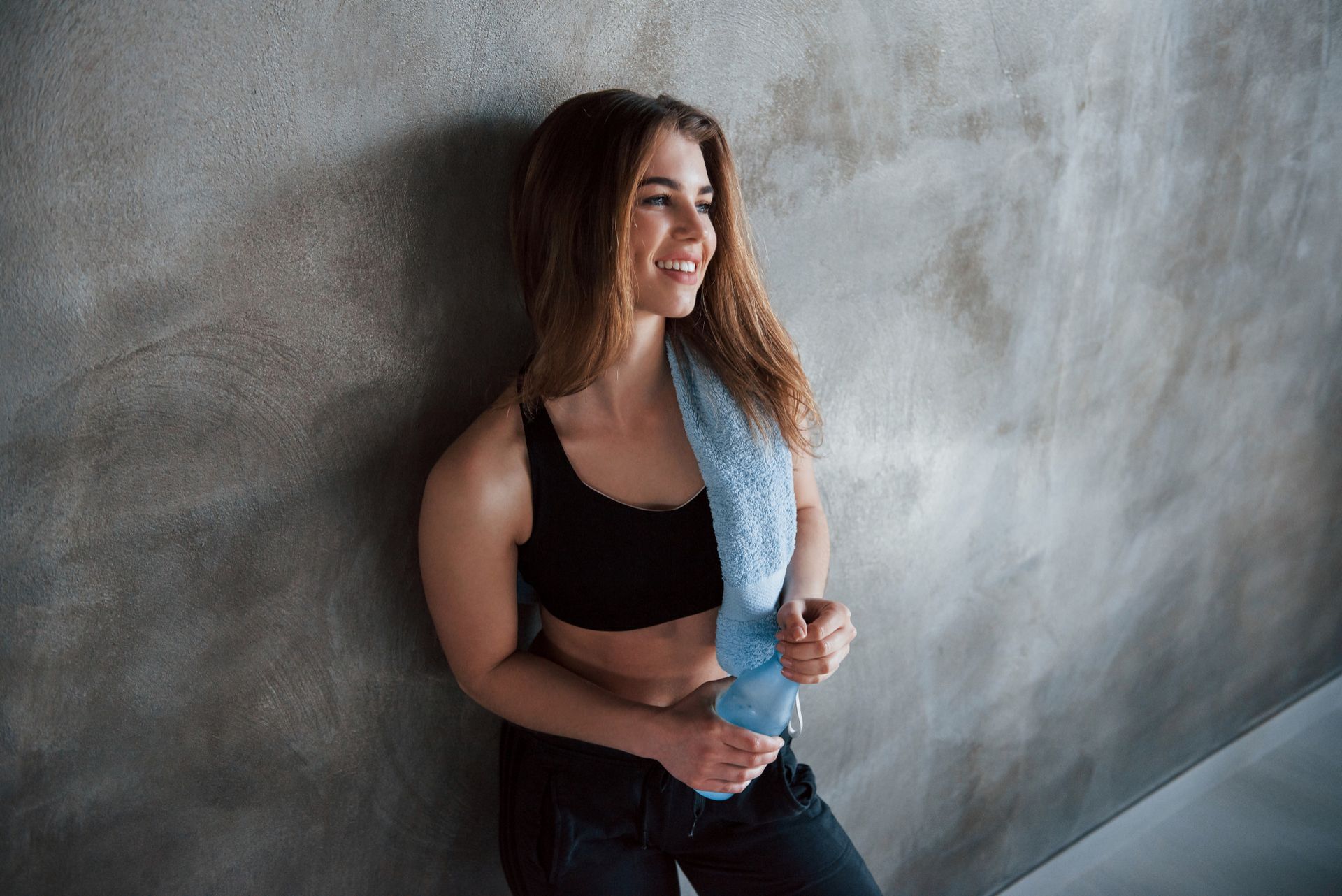 Woman in black sportswear leaning against a gray wall, holding a blue water bottle and smiling sideways.