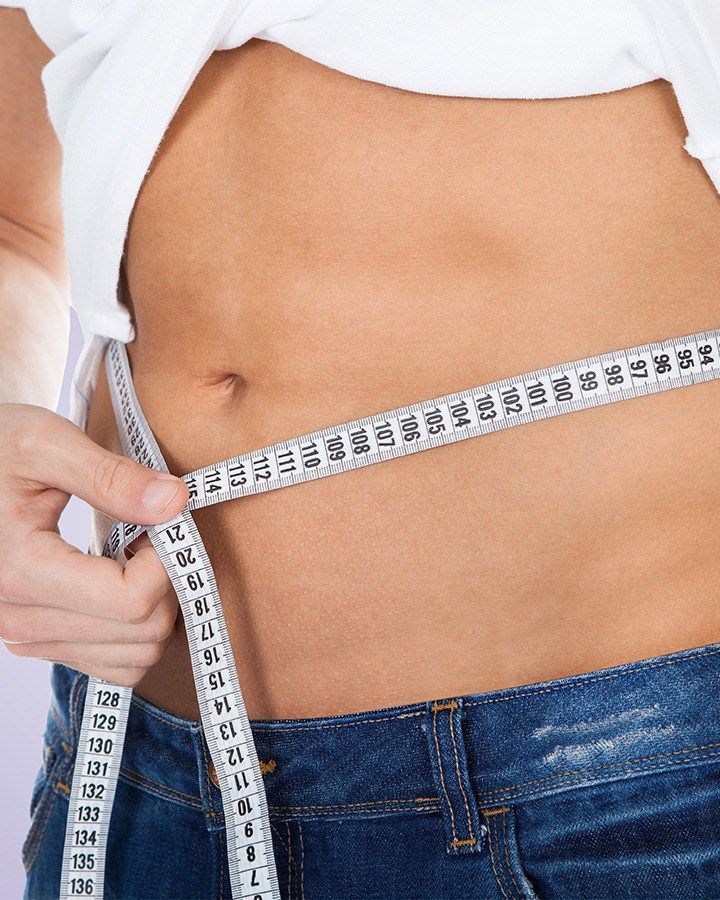 A person measuring their waist with a tape measure over blue jeans and a white shirt.