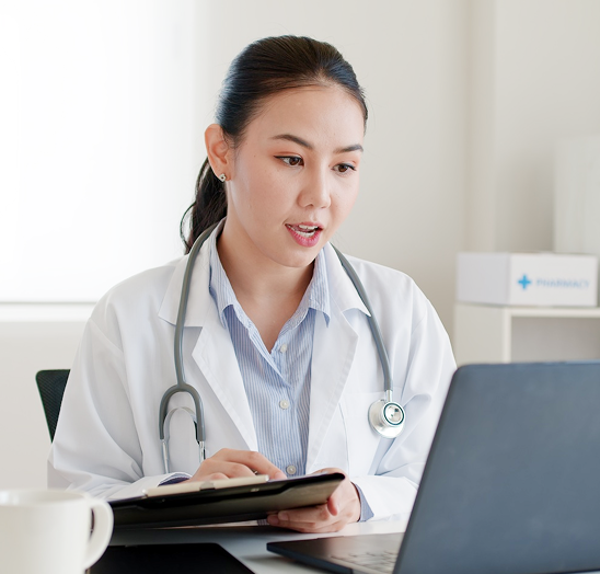 Doctor in white coat writing on a tablet beside a laptop in a bright office