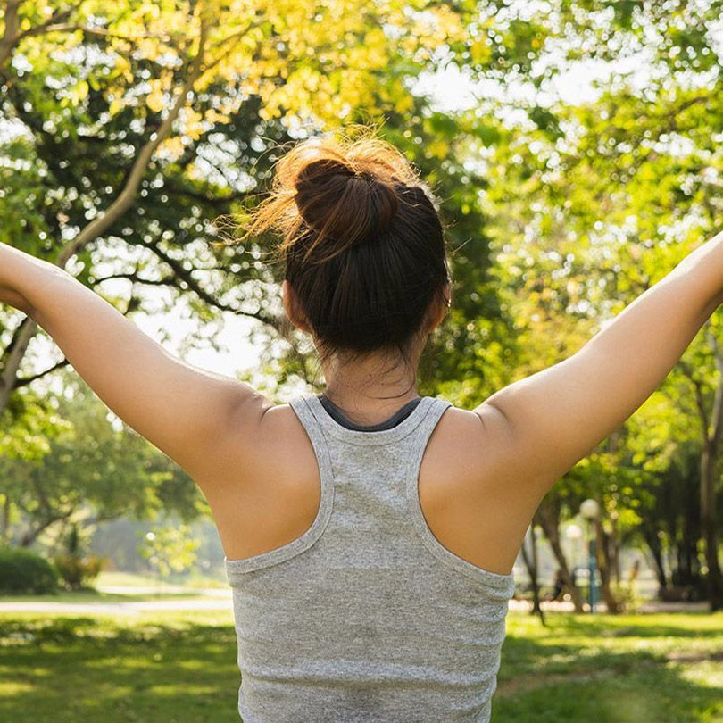 Person in a gray tank top stretching with arms raised in a sunny park