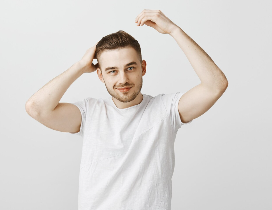 Man in a white T-shirt adjusting his hair against a plain light background