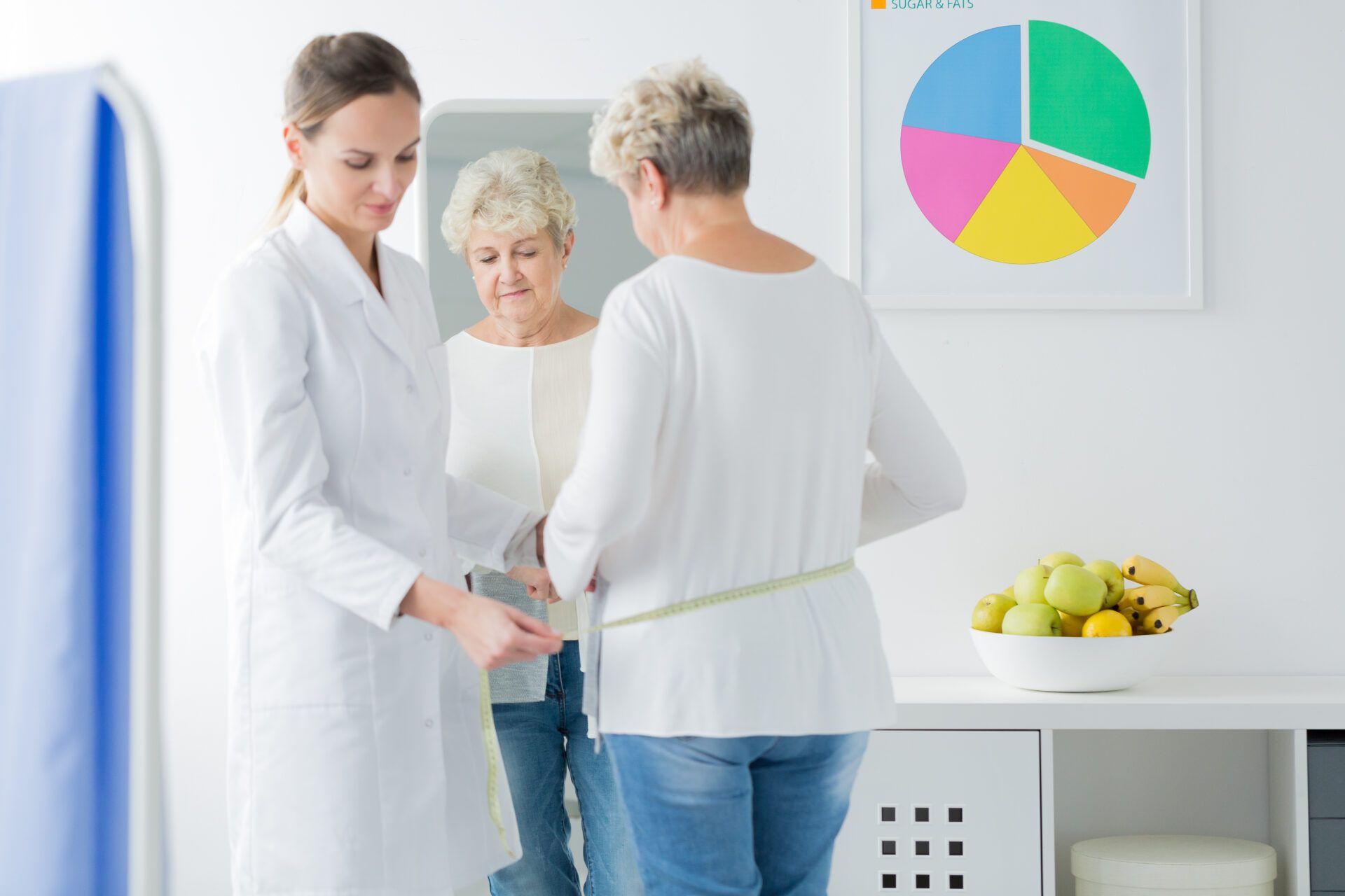 Doctor measuring a patient's waist in a bright clinic, with a health chart and fruit bowl nearby