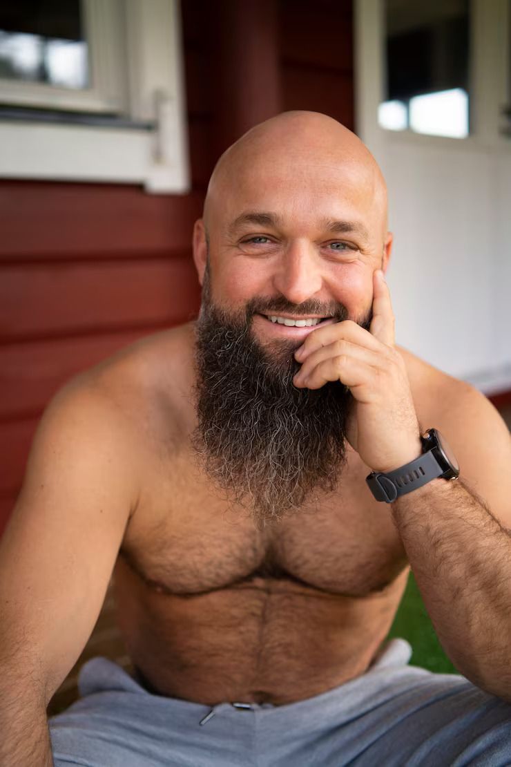 Smiling shirtless man with a long beard sitting on a porch, resting his hand on his face.