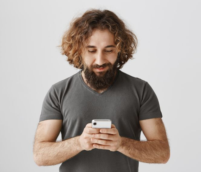 Man with curly hair and beard looking at a smartphone, wearing a gray T-shirt on a white background