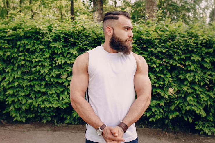 Man standing outdoors in a white sleeveless shirt in front of a green hedge, looking to the side.