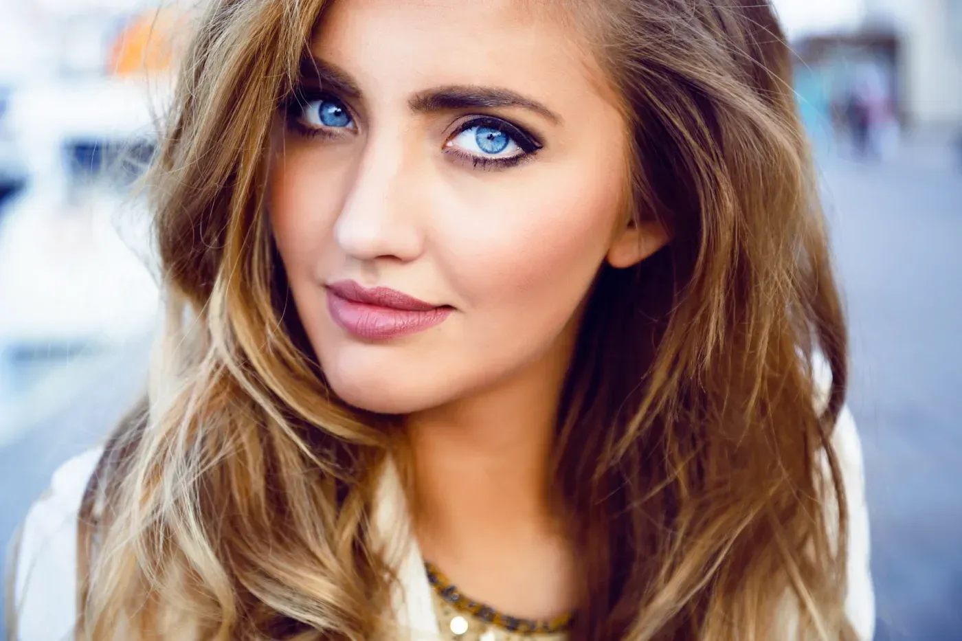 Close-up portrait of a woman with wavy blonde hair and blue eyes, wearing a light coat outdoors