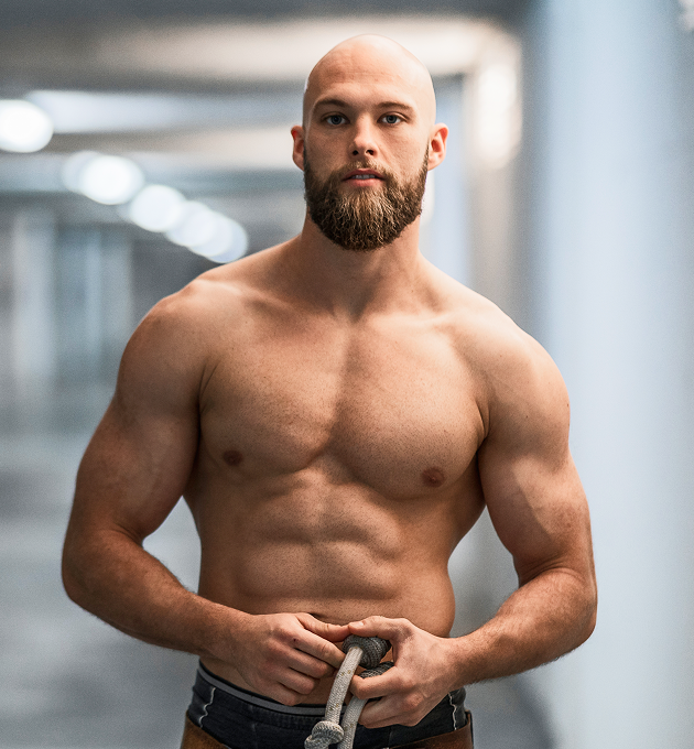 Shirtless muscular man standing in a gym, holding workout straps with a neutral expression.