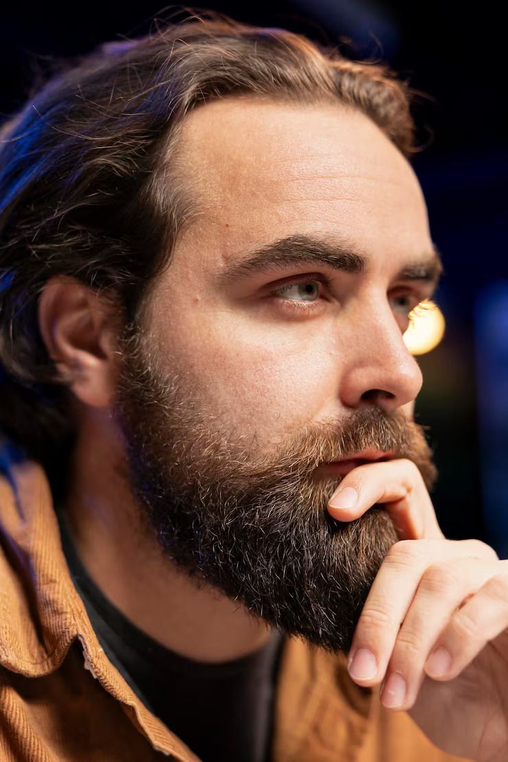 Close-up of a bearded person in profile, resting their hand on their chin in a dimly lit setting
