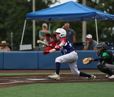 Youth baseball player in blue uniform bunts at home plate while catcher crouches nearby.