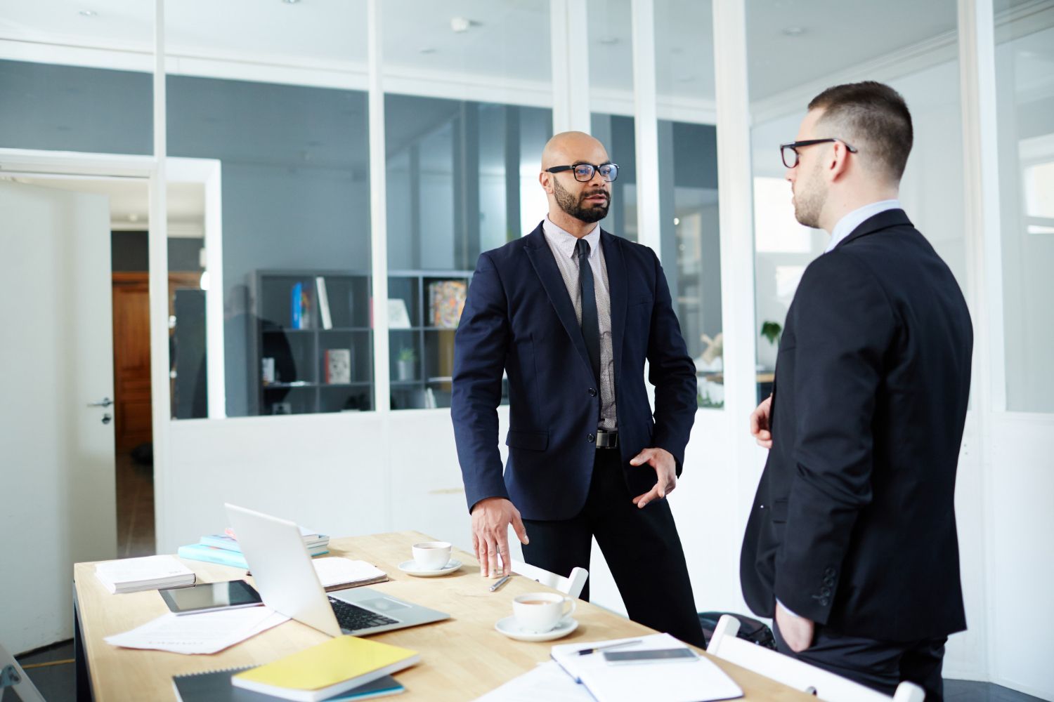 Two men in suits conversing in an office with a laptop, documents, and coffee on the desk.