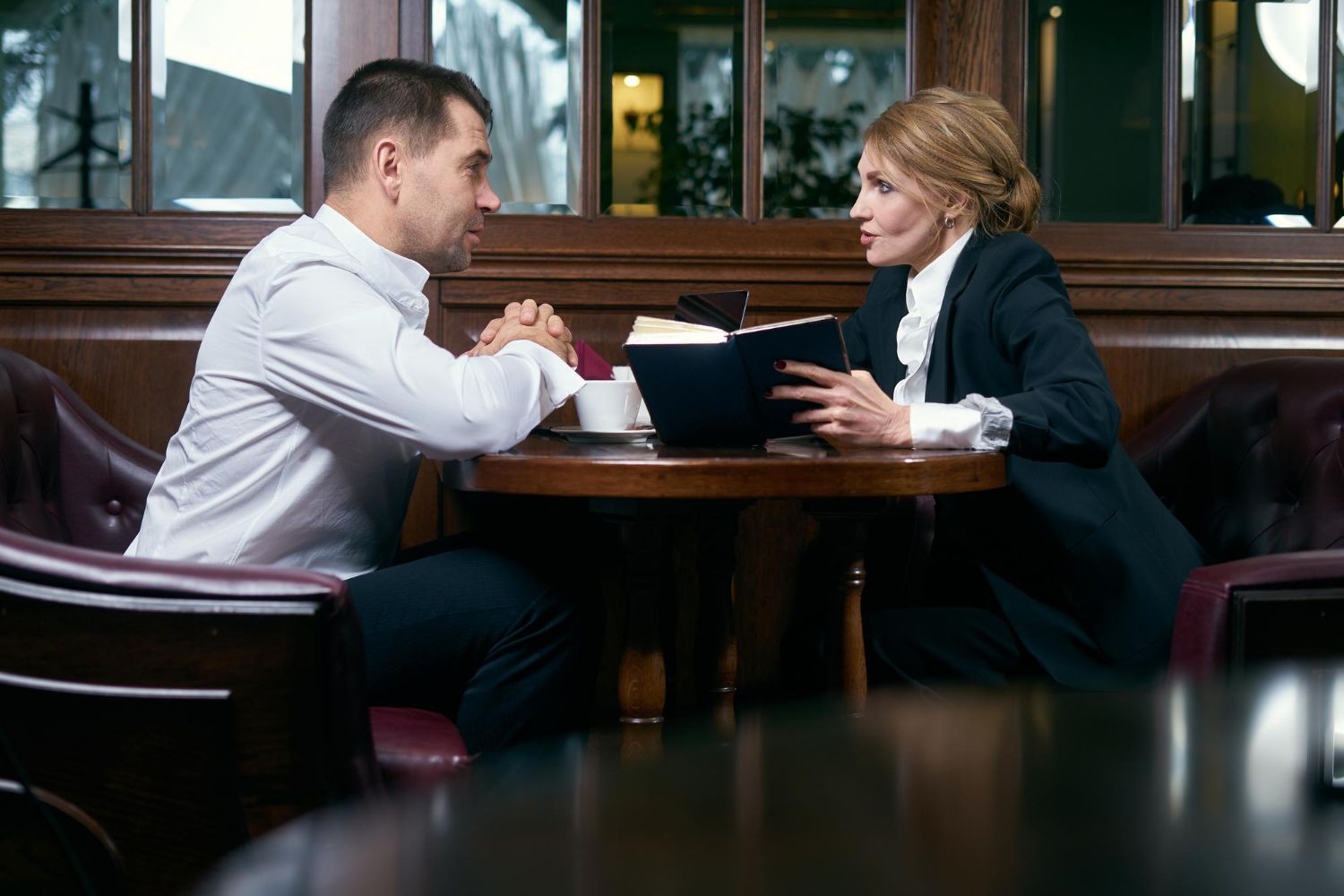 Man and woman at table in a restaurant, holding hands. Woman in suit reads from a notebook.