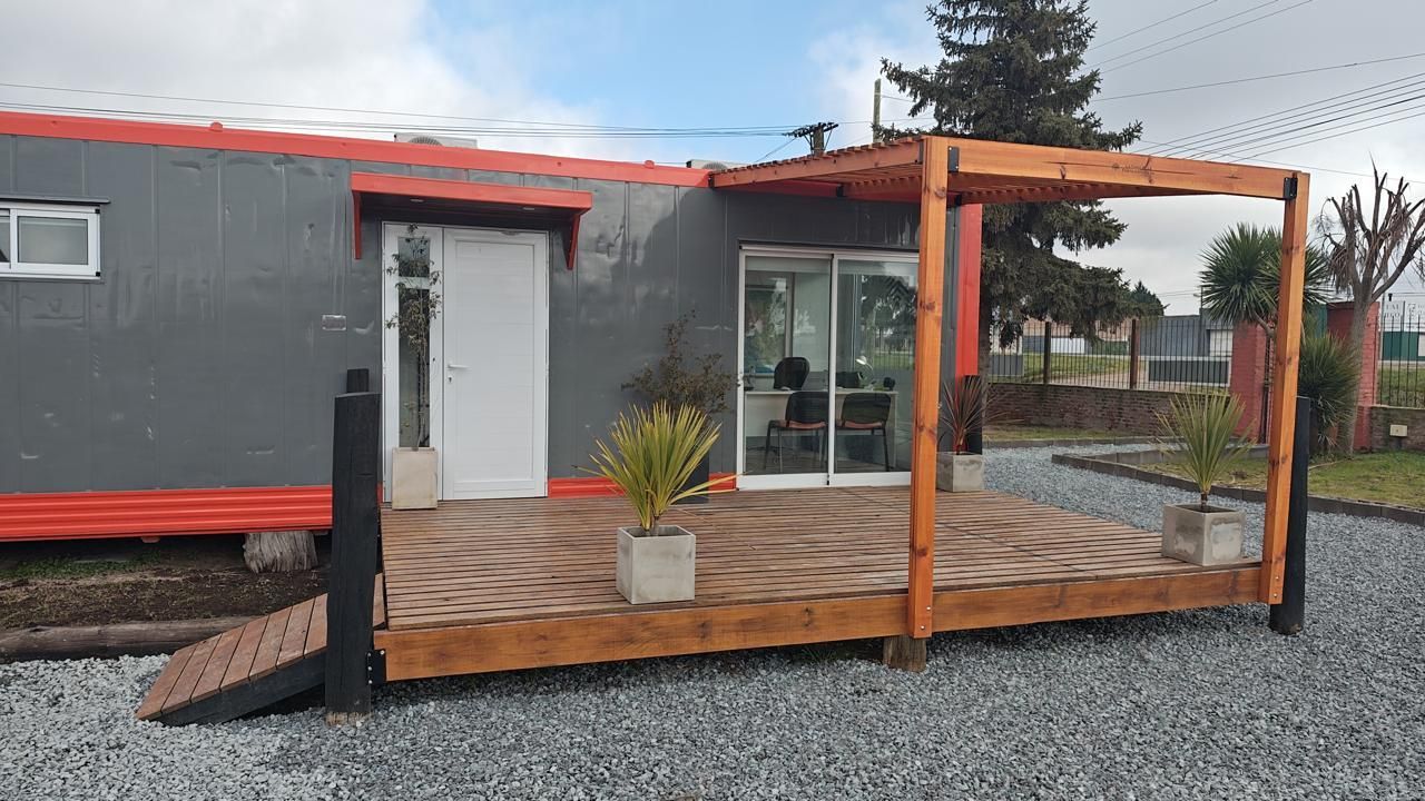 Edificio gris con deck de madera y pérgola; pequeñas plantas en deck; suelo de grava.