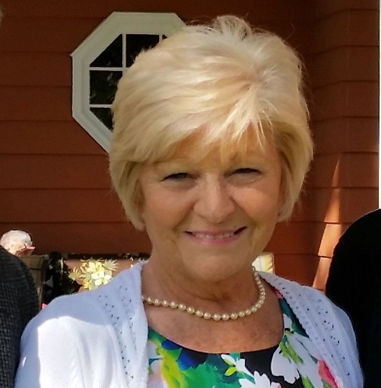 Smiling woman with short blonde hair, pearl necklace, and floral blouse standing outdoors by a house