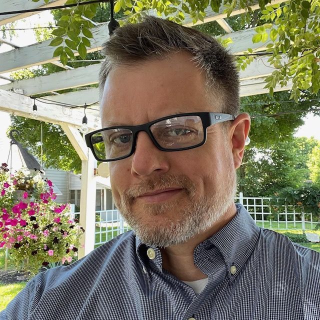 Smiling man with glasses in a garden under a pergola, with hanging flowers and greenery behind him