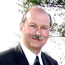 Smiling man in a dark suit and tie outdoors in front of leafy trees