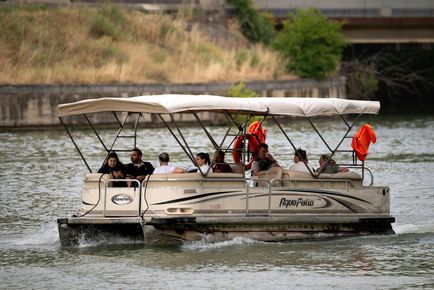 Pontoon boat carrying passengers on a lake, with people in orange jackets near the shore