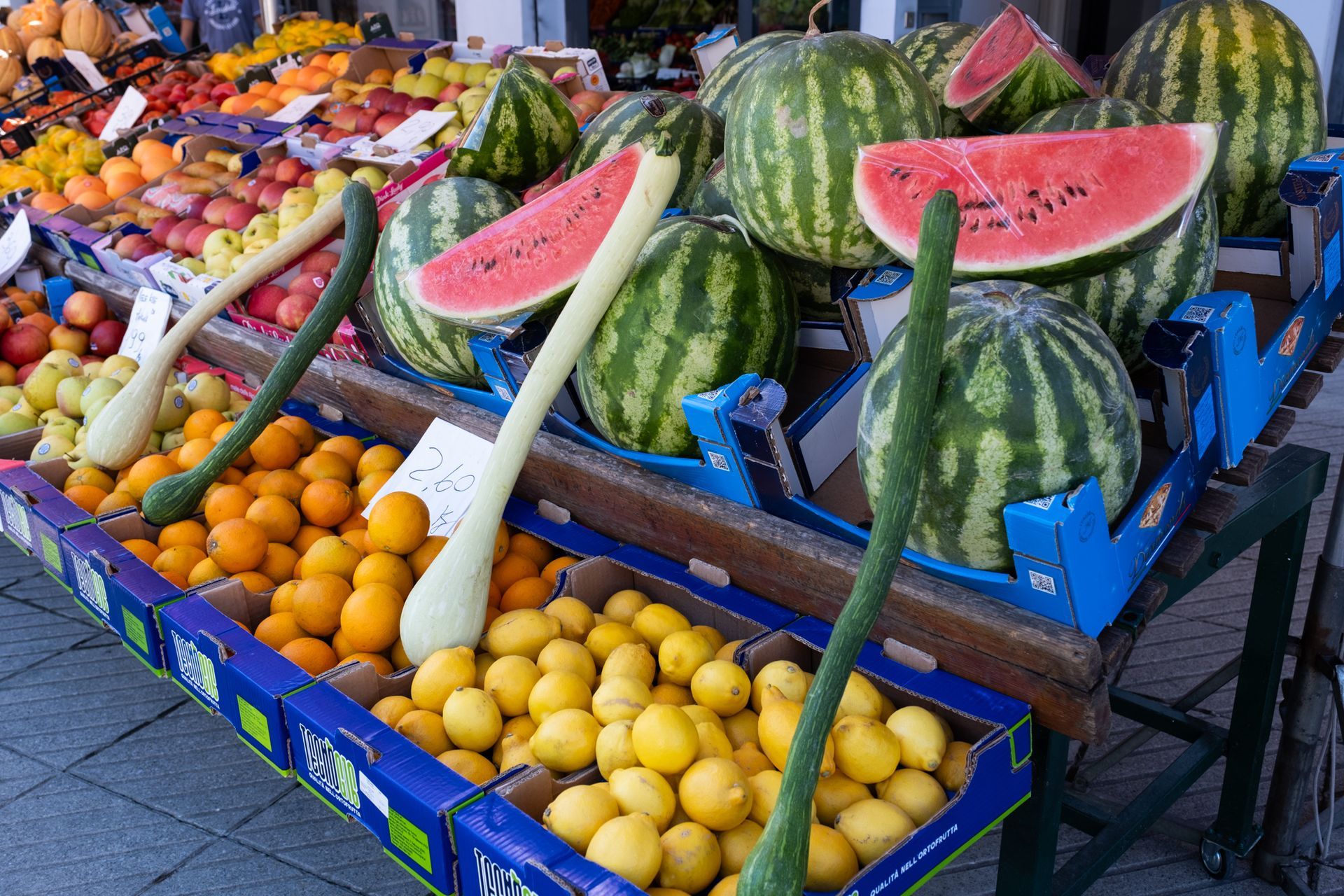 Banco di frutta colorato con angurie impilate, fette di melone, arance, limoni e prodotti assortiti in cassette.