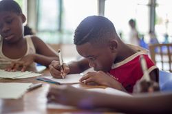 An elementary-age student in a red shirt is sitting at a table beside another student, writing on a piece of paper with a pencil as he bends his head down very close to the page.
