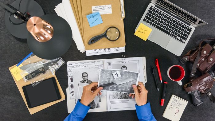 Top-down view of a desk with a laptop, notebook, camera, headphones, and hands reading a magazine.