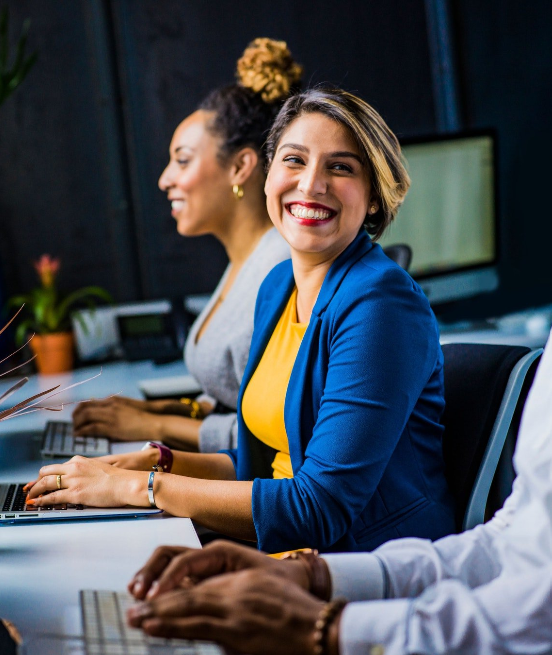Three coworkers in an office meeting, one woman smiling at the table in a blue blazer and yellow top.